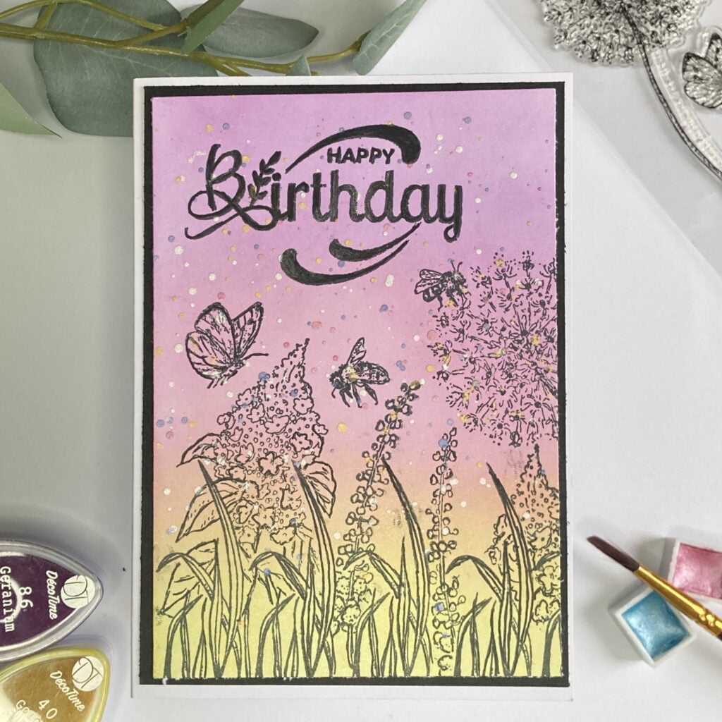 Full view of the handmade spring flower card, displayed upright. The card features a vibrant background with blended ink colors and metallic paint splatters. Delicate stamped flowers and grasses adorn the design, along with flying butterfly and bee stamps. A "Happy Birthday" sentiment is elegantly displayed in the center. Angle view of the spring flower card, showcasing its design and dimension. The card is displayed