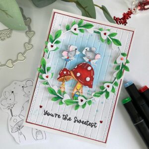Cute Handmade Valentine's Day card featuring adorable fairy mice within a wreath with hearts and leaves. The card is positioned at a unique angle, showcasing its charming design with vibrant leaves, red heart embellishments, and a dreamy blue background with embossed texture.