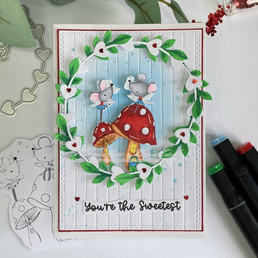 Featured image of a cute handmade Valentine's Day card with adorable fairy mice, surrounded by a frame/wreath with colorful leaves and red heart embellishments. The card is set against a dreamy blue background with embossed texture, showcasing creativity and love in crafting.