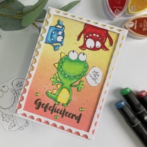Monteriffic handmade Monster Birthday card All Dressed Up Stamps 1 Top-down view of a handmade monster birthday card positioned diagonally on a surface. The card features three vibrant monster characters - a large green monster, a blue monster, and a red monster - with playful expressions. The background displays a coloruful gradient blending technique, adding depth and visual interest to the design.