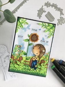 Little Sunflower scene card An angled top-down view of the entire handmade sunflower card, highlighting the detailed colouring of the sunflower girl, the layered background, and the carefully arranged die-cut elements. This image offers another perspective of the handmade sunflower card tutorial.
