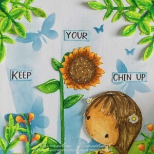 Close up picture featuring the sunflower and sentiment Keep Your Chin Up. A top-down view focusing on the upper portion of the handmade sunflower card. The "Keep Your Chin Up" text, positioned next to the sunflower, adds an uplifting message to the card. The vibrant colours create a cheerful atmosphere, showcasing another step in the handmade sunflower card tutorial.