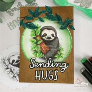 A top-down view of the completed handmade sloth card, laid flat. The card features a charming sloth image surrounded by branches and leaves against a textured background resembling tree bark. The sentiment "sending hugs" is elegantly displayed at the bottom of the card