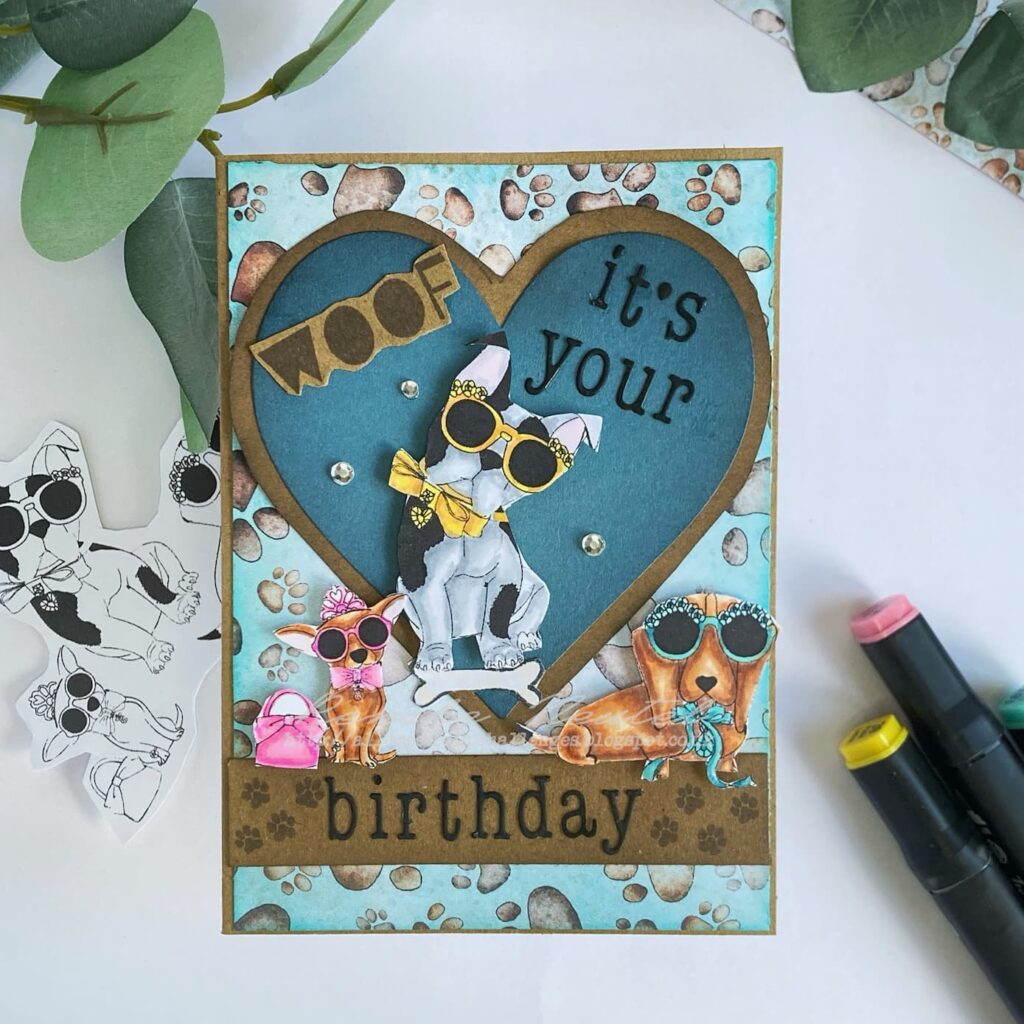 Handmade dog birthday card displayed flat and straight from above. The card features a background with blue paw print pattern paper, a layered heart in teal blue and kraft cardstock in the center, and three colored dogs with accessories. The sentiment reads 'Woof it's your birthday'