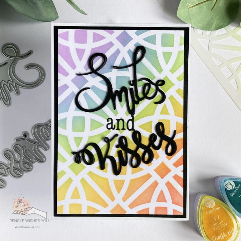 Top view of a handmade card featuring a rainbow gradient background and bold black die-cut sentiments 'smiles' and 'kisses.' The card is flat and positioned straight, showing the vibrant colors and elegant design."