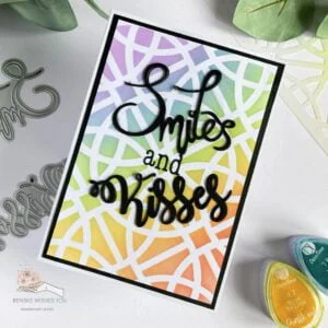 IMG_6867-(W) Top view of a handmade card with a rainbow gradient background and bold black die-cut sentiments 'smiles' and 'kisses.' The card is positioned at an angle, highlighting the smooth blending of colors and the striking contrast of the black sentiments.