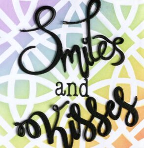 Smiles and Kisses Close-up of the bold black die-cut sentiments 'smiles' and 'kisses' on a handmade card with a rainbow gradient background. The detailed texture and dimension of the die-cut words are clearly visible.