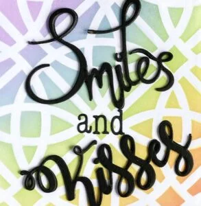 Smiles and Kisses Close-up of the bold black die-cut sentiments 'smiles' and 'kisses' on a handmade card with a rainbow gradient background. The detailed texture and dimension of the die-cut words are clearly visible.