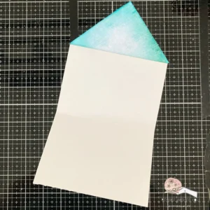 Envelop flap card folded open pen view of the base of the envelope flap card, with the flap attached but undecorated. The photo shows the construction of the card, highlighting the folding technique used to create the envelope flap closure. The card base is made from white cardstock, with visible creases where the folds are, preparing it for the addition of patterned paper and embellishments