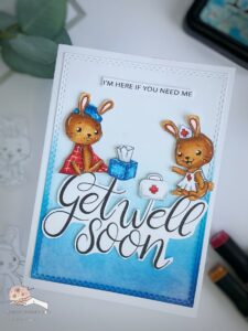 Get Well Soon Card Isabelchristina Handmade get well soon card positioned at an angle, depicting two cute bunnies — one sick and one as a nurse — along with a bold 'Get Well Soon' sentiment and decorative elements like a tissue box and a doctor's bag, surrounded by crafting tools and supplies such as alcohol markers and distress inks.