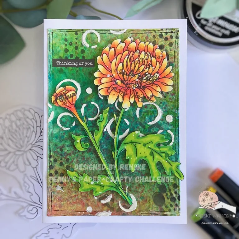 Top-down view of a handmade mixed media flower card lying flat and centered. The card features a brightly colored chrysanthemum in shades of yellow, orange, and red, placed on a textured, layered background created with acrylic sprays and stencils. A sentiment in the top left reads 'Thinking of You.