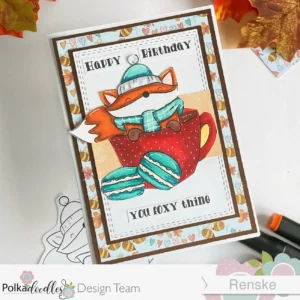 Autumn birthday card hot chocolate fox Polkadoodles n angled overhead view of the autumn birthday card, displaying its layered design. The card features a playful arrangement of a fox in a hot chocolate cup, surrounded by autumn-themed elements like leaves and macarons. The layers include a light blue patterned paper with hearts, a rich brown cardstock layer, and a white rectangle die-cut panel that adds dimension. The card has a cheerful and inviting autumn aesthetic.