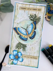 Butterfly greeting card with vintage style (2) Top-down angled view of a vintage butterfly greeting card, showing a distressed wooden background with layered details. The card showcases a brightly coloured butterfly, a blue flower accent, vintage clock and leaf die-cuts, and distressed edges, all arranged on a slimline card with subtle vintage-inspired textures.