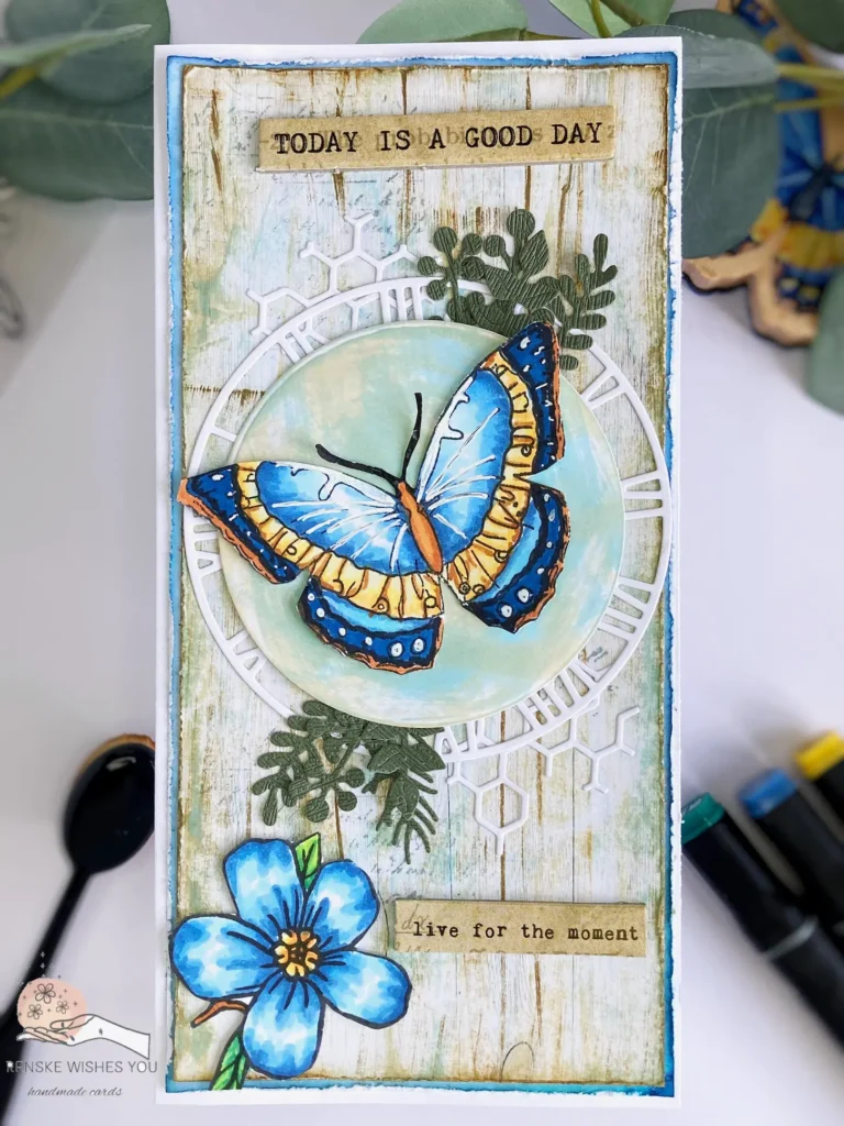 Top-down view of a vintage butterfly greeting card. The card features a distressed wooden background, a vibrant die-cut butterfly in soft blues and yellows, a blue flower accent, and vintage clock and leaf embellishments, all arranged on a slimline card with subtle layers for added texture