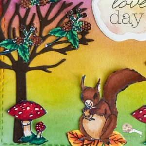 Close up picture of the squirrel sitting on leaves nest to a tree and mushrooms "Close-up of squirrel, tree, and mushroom on a handmade autumn card. The squirrel sits on colorful autumn leaves beside a tree and a red mushroom, creating a woodland scene. The ink-blended background enhances the warm autumn theme of the card