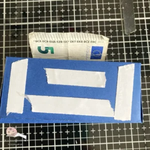 Double sided tape to attach the background to the box Back view of the DIY money roll box, showing double-sided tape on the bottom flaps. The tape will secure the background to the box once it's assembled. Tutorial step for creating a money roll box.