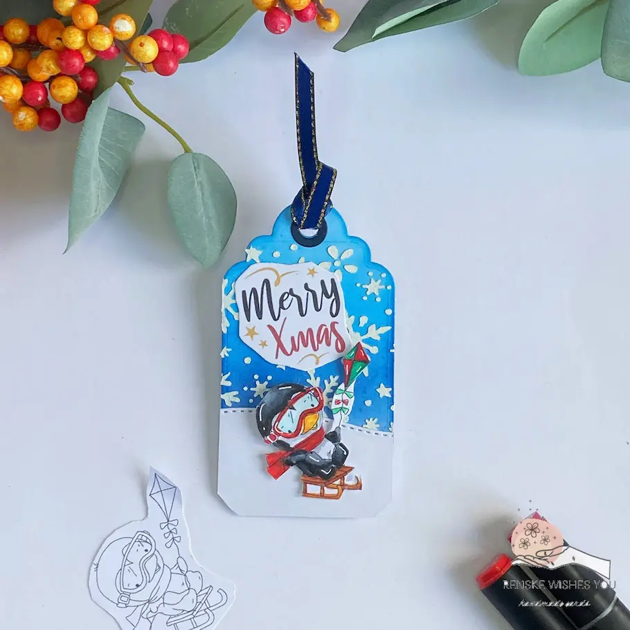 Finished penguin christmas tag with waddy on a sleight Polkadoodles Overhead shot of a penguin tag featuring Waddy Penguin on a sleigh. The vibrant background and snowy hill create a charming winter scene, with dimension added by foam tape for a standout design.
