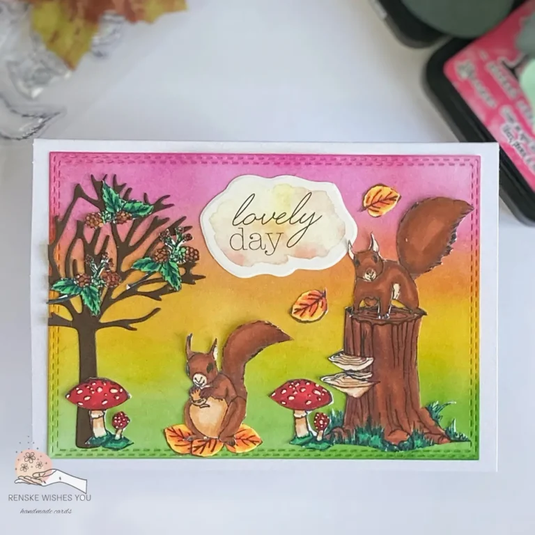 Handmade autumn card with squirrels and ink blending – top-down view. The card features a warm, autumn-themed design with a playful squirrel on a tree stump, and a squirrel sitting on autumn leaves surrounded by fall leaves, mushrooms, and trees. The background is subtly blended with pink, yellow and green distress ink to create a soft autumn atmosphere