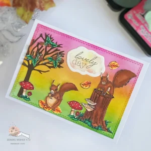 Handmade autumn card with squirrels and ink blending Marianne Deisign 3 Handmade autumn card with squirrels and ink blending – angled view. A slightly angled top-down view of the autumn card highlights a charming squirrel on a tree stump, and a squirrel on autumn leaves surrounded by colourful fall leaves, mushrooms, and trees set against an ink-blended autumn landscape. This perspective showcases the card's distress ink background with pink, yellow and green colours.