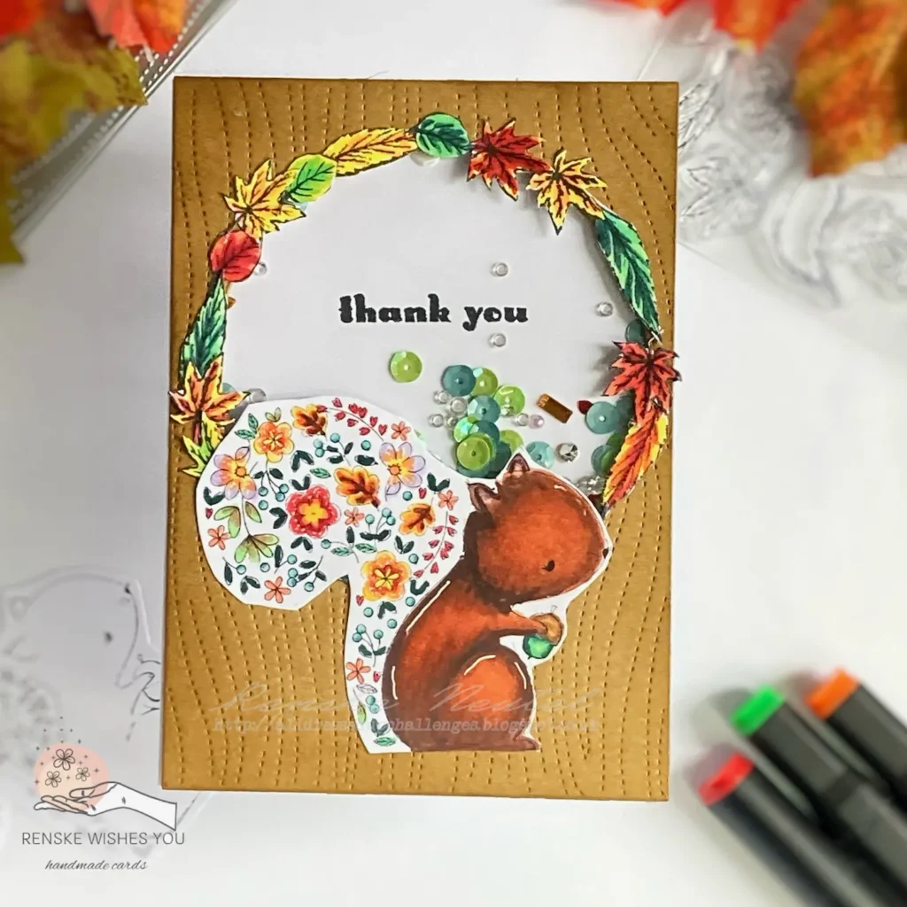 Top view of a completed autumn shaker card featuring a squirrel holding an acorn. The squirrel's tail is made up of leaves, flowers, berries, and hearts. The card has a shaker window with autumn-themed elements inside and a thank you sentiment. Perfect DIY for a handmade autumn shaker card.
