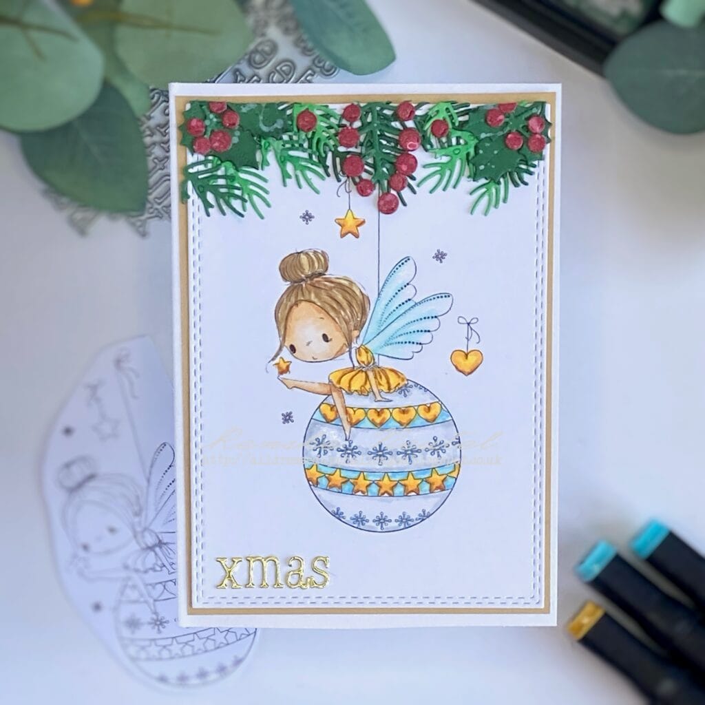 Top-down view of the completed fairy Christmas card, showing the centred Fairy Bauble digital stamp coloured in festive tones, surrounded by intricate holly, conifer branches, and berries. The card’s layout is balanced and festive, with soft shimmer accents and die-cut details enhancing the seasonal design