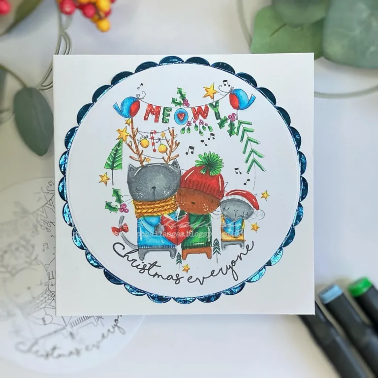 op-down view of a handmade Meowy Christmas card, featuring a festive digital stamp of three cats wearing holiday attire surrounded by a decorative metallic blue border. The card base is white, and the digital stamp showcases vibrant colouring with alcohol markers. Perfect for holiday crafting inspiration.
