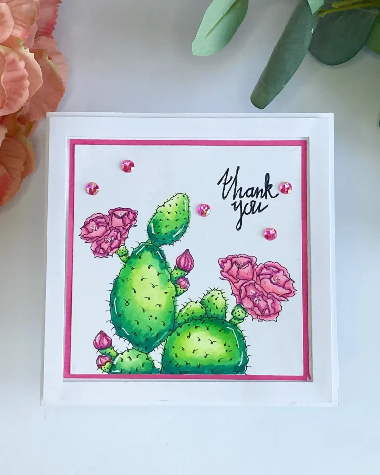 Top-down view of the finished handmade cactus thank you card. The card features a hand-colored cactus illustration framed with a pink border, a white die-cut frame, and pearl embellishments on a white card base