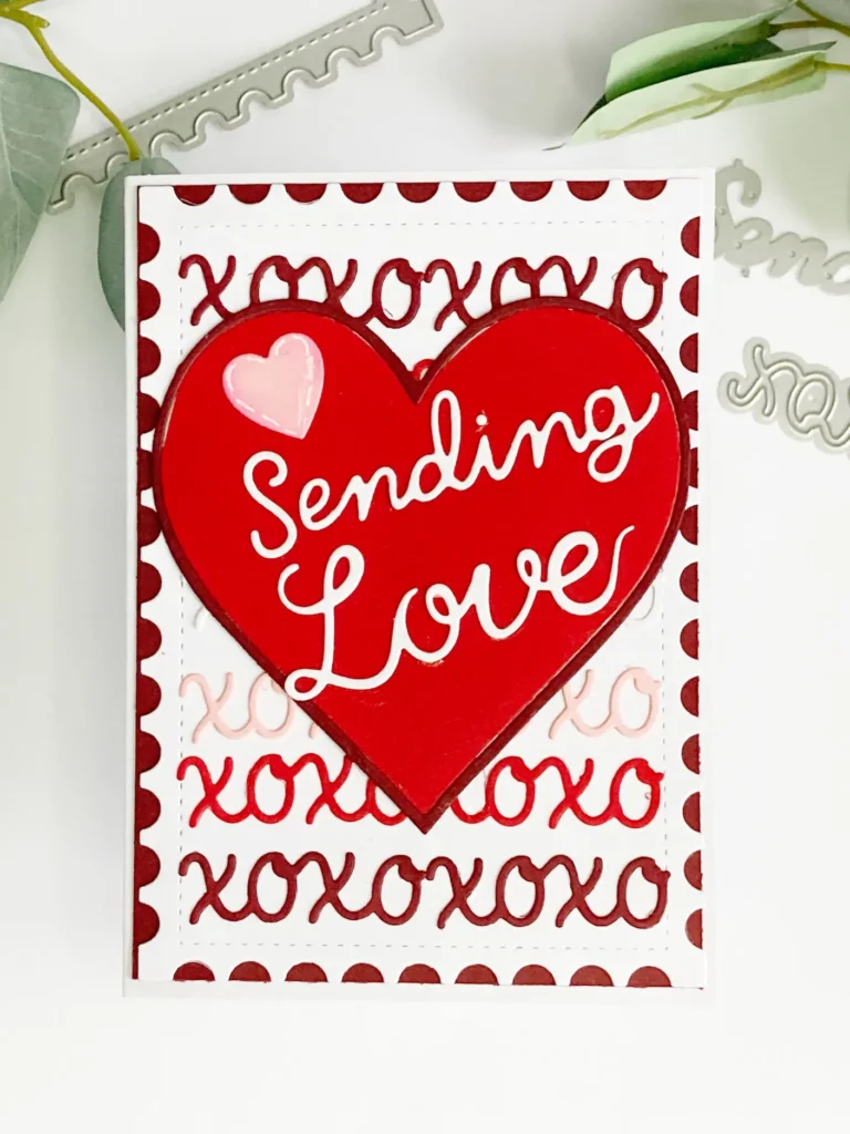A top-down view of a completed DIY Valentine card design featuring a layered xoxo card background in red, pink, and white, with a central heart detail that reads “Sending Love.”