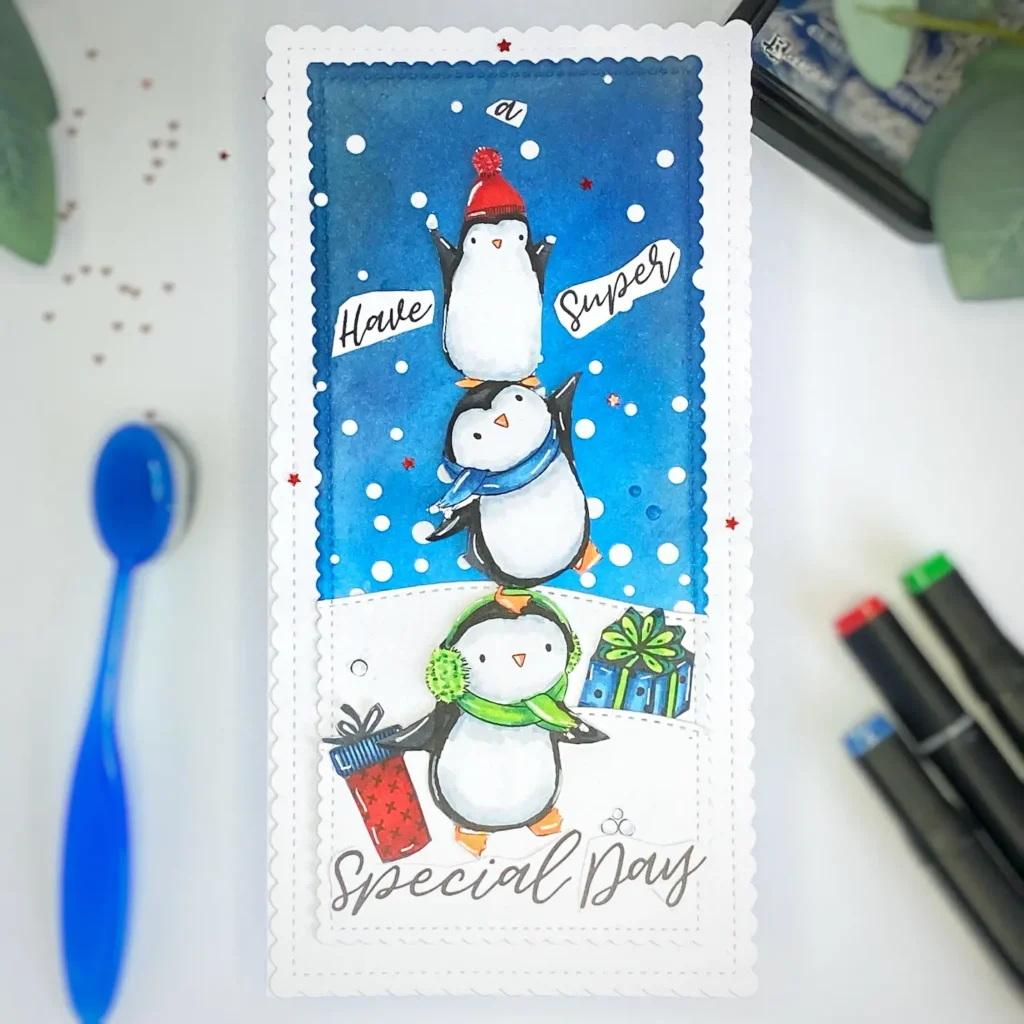 Top-down view of a handmade Penguin Birthday Card featuring three stacked penguins, snowdrifts, and colourful gift stamps. The card is displayed on a night sky background, showing its playful winter theme.