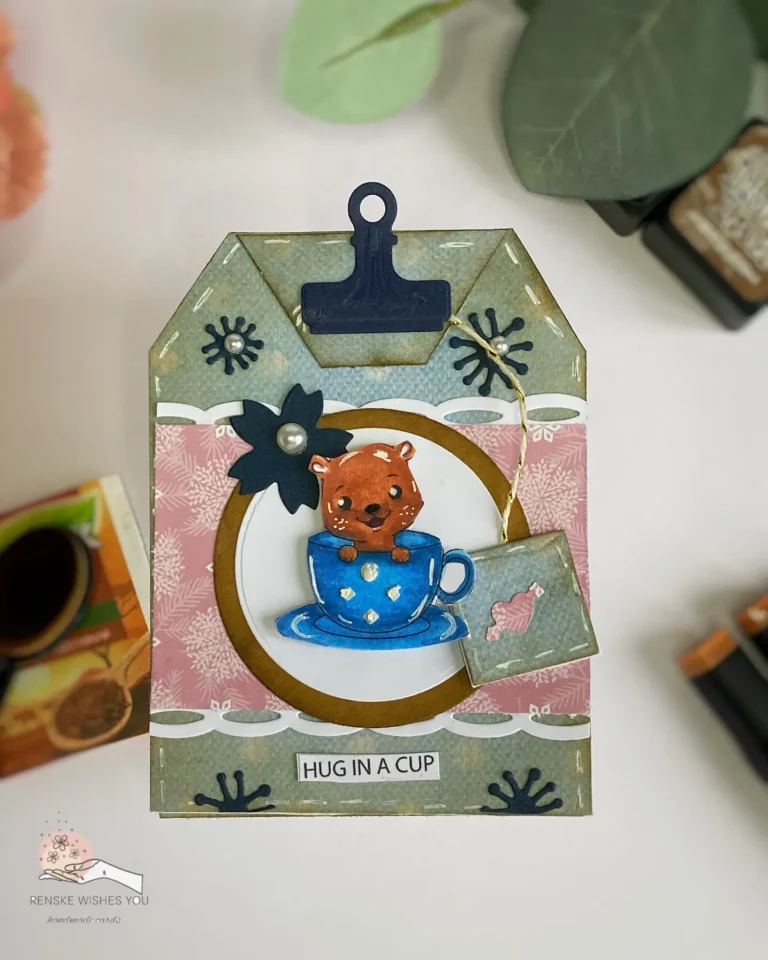 Top-down view of the shaped tea bag card showing its detailed front design. The card features a cozy pink and blue color palette, intricate layers, and the sentiment "Hug in a Cup" prominently displayed.The centre piece is a bear in a tea cup.