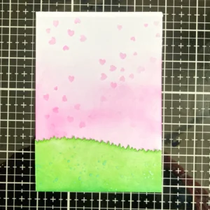 Finished background A view of the completed background for the handmade Mother's Day card. The soft pink blended base with a heart stencil pattern is now fully dried and ready for assembly. The die-cut grass is attached at the bottom, completing the layered backdrop for the card’s main elements.