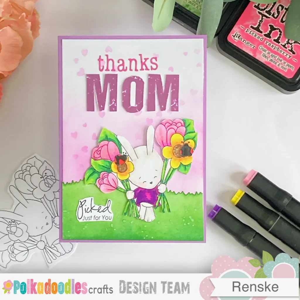 top-down view of the handmade Mother's Day card, featuring a pastel purple card base with a soft pink blended background. The focal point is a cute bunny holding a bouquet of flowers, surrounded by a stencilled heart pattern. A die-cut grass border at the bottom adds texture, and the "Thanks Mom" sentiment is displayed in ourple and pink lettering