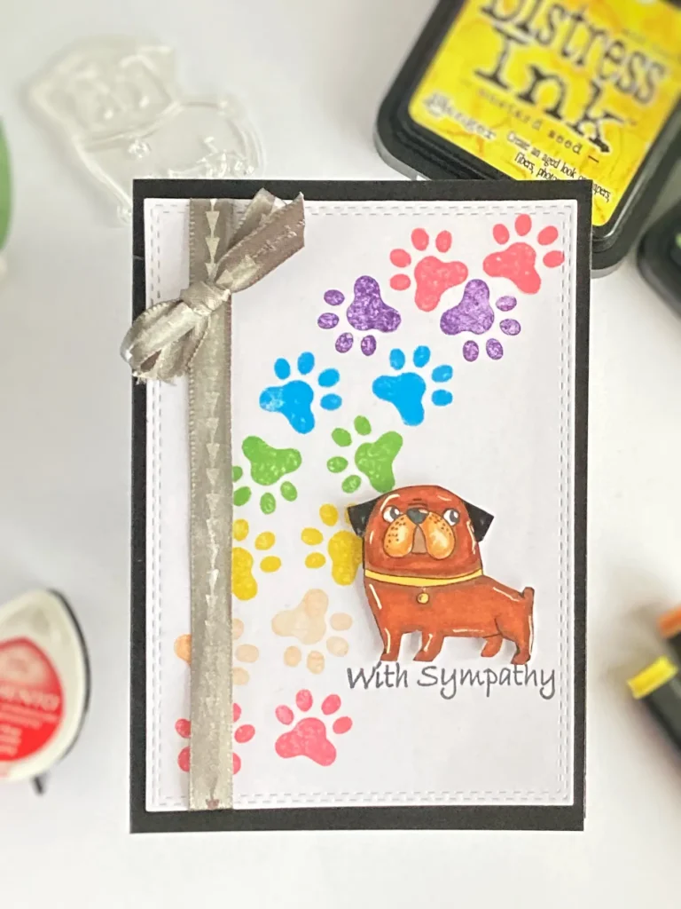 Handmade pet sympathy card captured from a top-down view, lying straight. The card features a rainbow paw print background, a stamped and coloured dog image, and a heartfelt "With Sympathy" message.