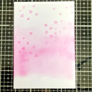 Ink blended background pink and hearts A close-up of the background created for the handmade Mother's Day card. The Distress Ink blending transitions from a rich pink at the bottom to a lighter shade at the top. A heart stencil was used to add soft, floating heart patterns, giving the background a subtle yet charming texture.