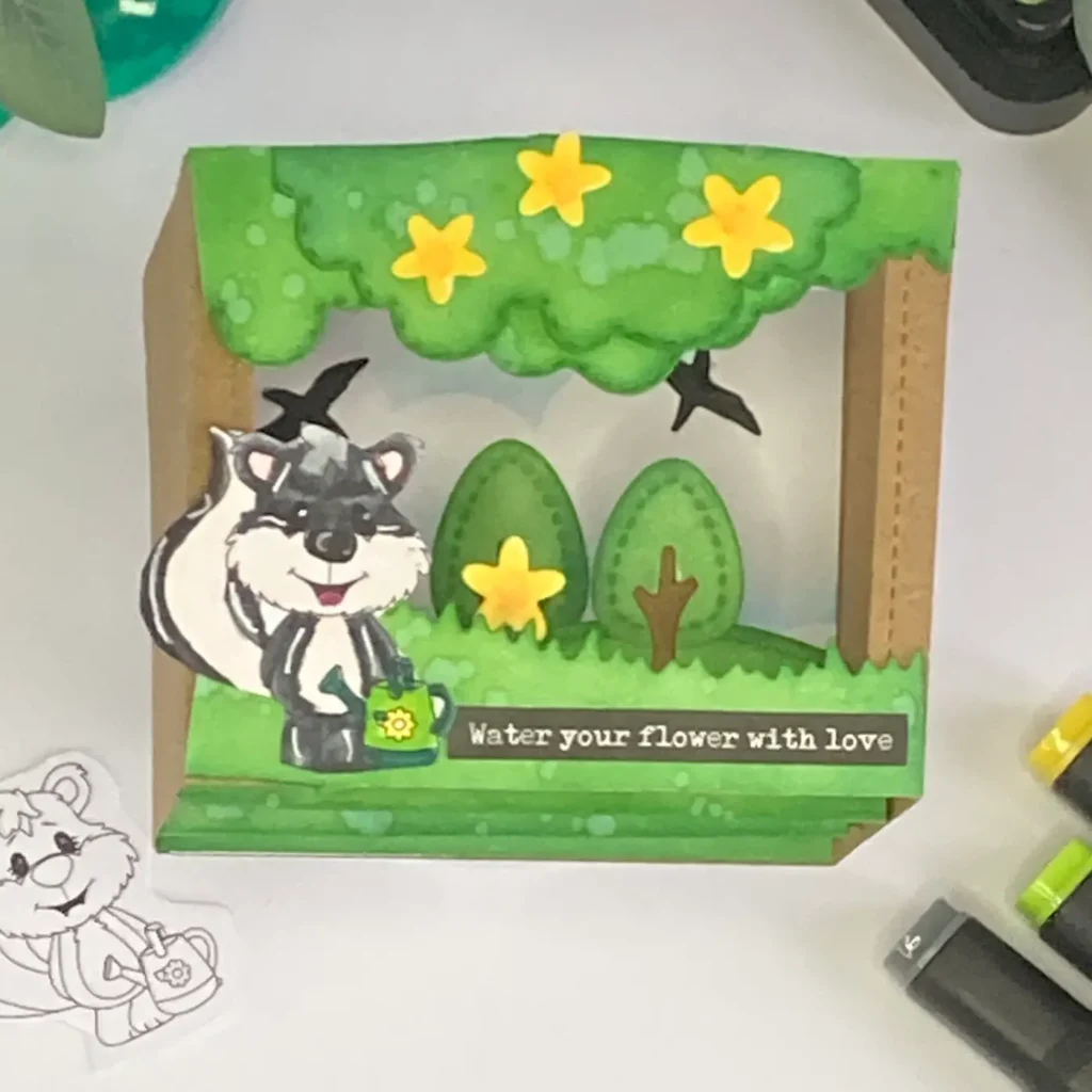 Full view of the finished card from the shadow box card tutorial. The skunk holding a watering can is surrounded by layered trees, grass, and flowers, creating a dimensional scene inside the box frame.