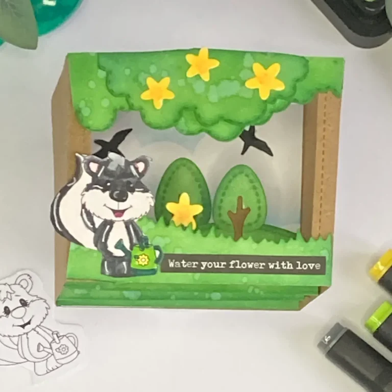 Full view of the finished card from the shadow box card tutorial. The skunk holding a watering can is surrounded by layered trees, grass, and flowers, creating a dimensional scene inside the box frame.