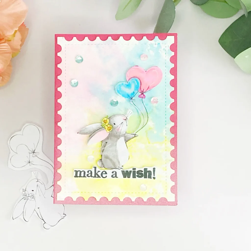 A handmade bunny card featuring a soft ink-smooshed background in shades of blue, pink, and yellow. The centre showcases a coloured digital stamp of a bunny holding pastel balloons. The sentiment "Make a Wish" is stamped in black on a white strip at the bottom. Small embellishments in coordinating colours add balance to the design