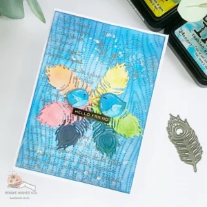 Mixed Media Bird and Feather card (2) Top-down angled view of the mixed media card, highlighting its dimensional elements. The feather wreath encircles two birds, with ink splatters and script stamping adding artistic detail. The layered background consists of soft blended tones, stencils, and paste. Light catches the raised textures, emphasizing the depth created by mixed media techniques. This mixed media card tutorial explores how to combine stamping, stencilling, and layering for a unique effect.