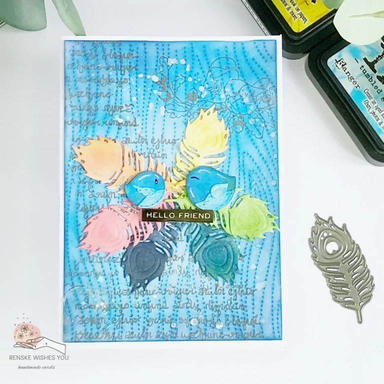 Top-down view of a mixed media card lying flat. The design features a central wreath of layered feathers in rainbox colours, creating a textured frame. Inside the wreath, two illustrated birds perch closely together, surrounded by delicate ink splatters and subtle script stamping. The background has a distressed effect with blended colours and stencilled patterns, adding depth. This mixed media card tutorial showcases various techniques for texture and layering