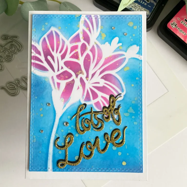 Overhead view of a handmade spring flower card placed upright, showing a bold pink and purple stencilled flower in the centre with a soft blue ink-blended background. The gold foiled “Lots of Love” sentiment is positioned diagonally, and flat-back pearls are added in opposite corners for balance.