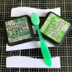 Grass strips with distress ink Die-cut grass strips inked with Distress Ink in Mowed Lawn and Pine Needles. The ink pads and blending brushes are visible next to the strips, showing the two-tone effect used for the grassy areas in the tunnel card.