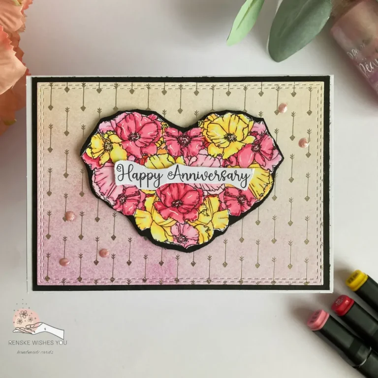 Overhead view of a handmade anniversary card lying flat. The card features a floral heart in soft pinks, yellows, and reds, outlined in black and centred on a soft gradient background with silver foil arrows. The card has clean black matting and a white base, creating a modern yet romantic look.