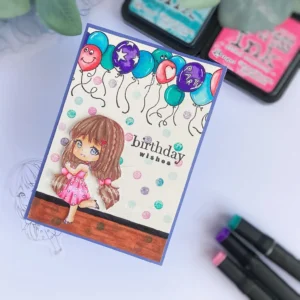Handmade birthday card with festive balloons Angled top-down photo of the handmade birthday card highlighting the foam-mounted coloured girl, confetti accents, and the dimension of the layered background.