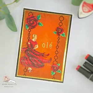 Handmade flamenco card Sheepski Designs (2) Angled top-down view of the flamenco dancing card featured in the tutorial. The rich orange background shows a soft stencilled pattern, framed by golden foil and layered onto a white card base. The dancer image pops with dimension, wearing a flowing red dress that swirls to the side. Three large red roses with green leaves accent the black die-cut chain borders. The gold “olé” sentiment sits to the right of the dancer, creating a bold focal point.