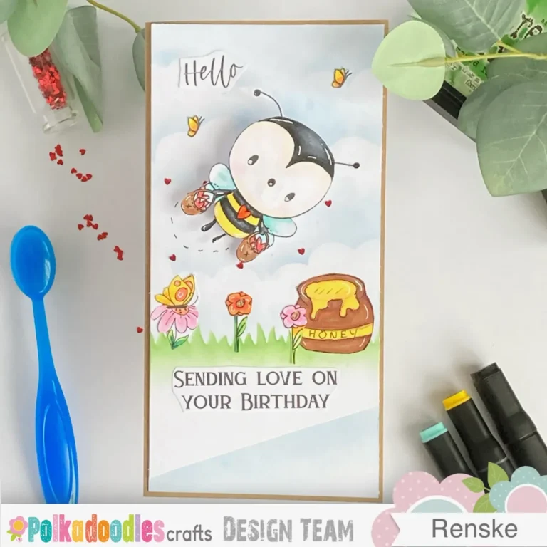 Top-down view of the finished bee birthday card featuring a wobble element, layered slimline background with stencilled grass and clouds, and a glittered bee holding heart baskets.