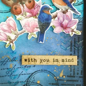 Close up of the sentiment Close-up of the sentiment ‘with you in my mind’ on a mixed-media card with paper die-cuts, placed over an inked and textured background with subtle stamping