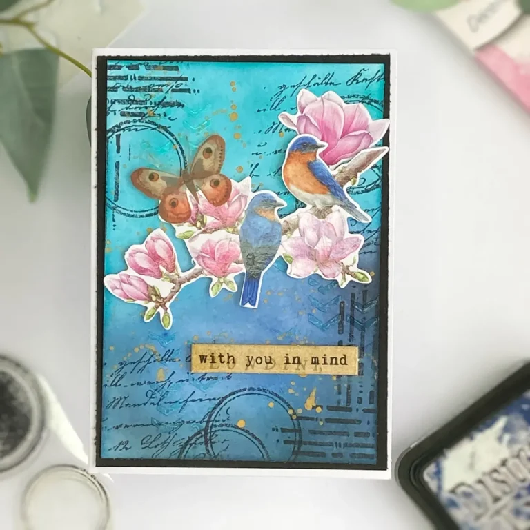 Top view of a finished mixed-media card with paper die-cuts featuring two birds on a branch, layered flowers, stamped background, and blended distress ink in soft blues and greens