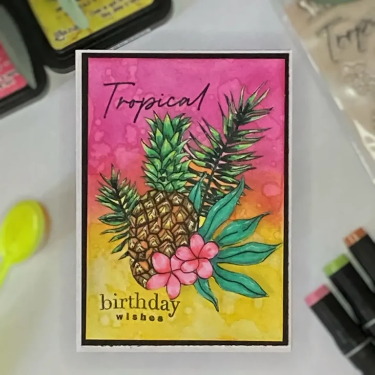 Handmade tropical birthday card photographed from above, showing a bright ink-blended background with a coloured pineapple stamp, tropical flowers, and bold sentiment.