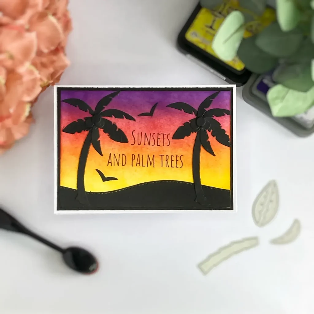Handmade sunset card with bold black palm tree silhouettes, blended ink background in warm yellow, orange, red, and purple tones, shown from above.