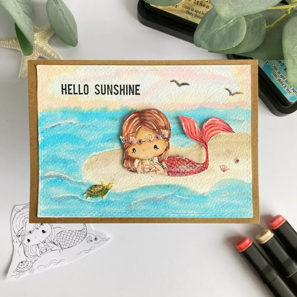 "Top-down view of a finished handmade mermaid card, showing the mermaid resting on a sandy bank with a flowing watercolour ocean and a tiny turtle nearby, surrounded by summer-themed details.